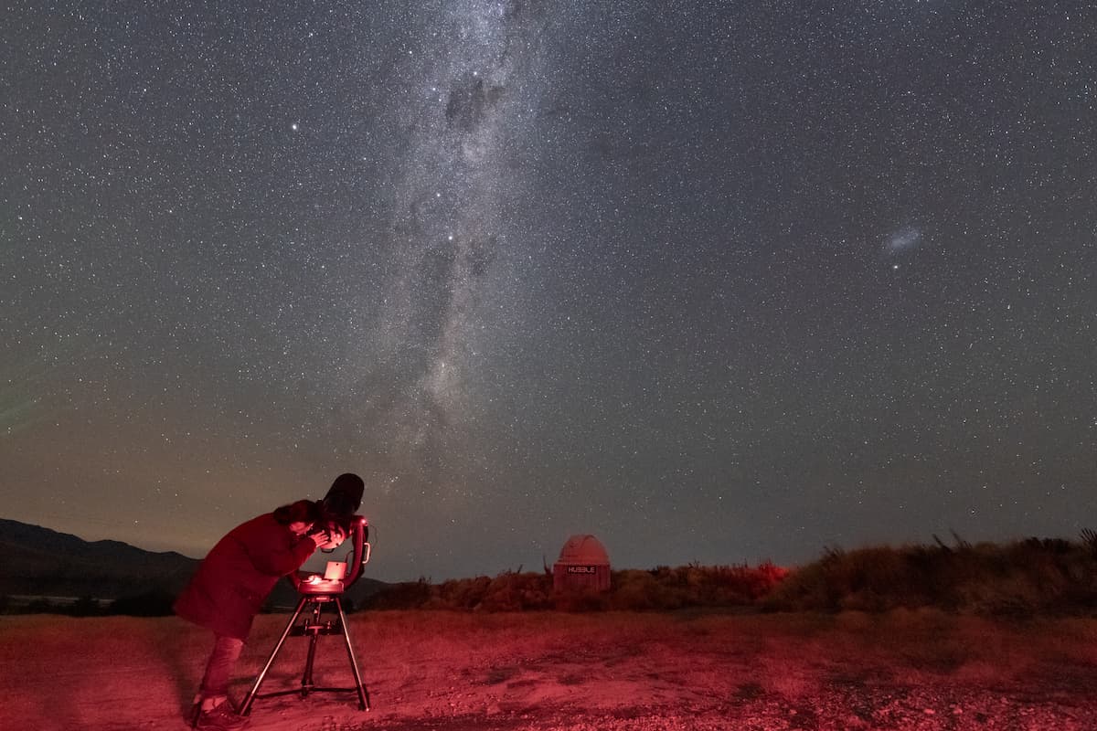  The image shows a person looking through a telescope under a clear, star-filled night sky. The Milky Way galaxy is vividly visible, stretching across the sky with its bright cosmic clouds. The person is standing in an open field, illuminated by a soft re