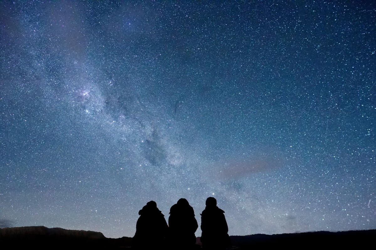 The image shows three silhouetted figures seated outdoors at night, gazing up at a stunning star-filled sky. The Milky Way is prominently visible, stretching across the sky with bright clusters of stars and cosmic dust. The silhouettes add a sense of scal